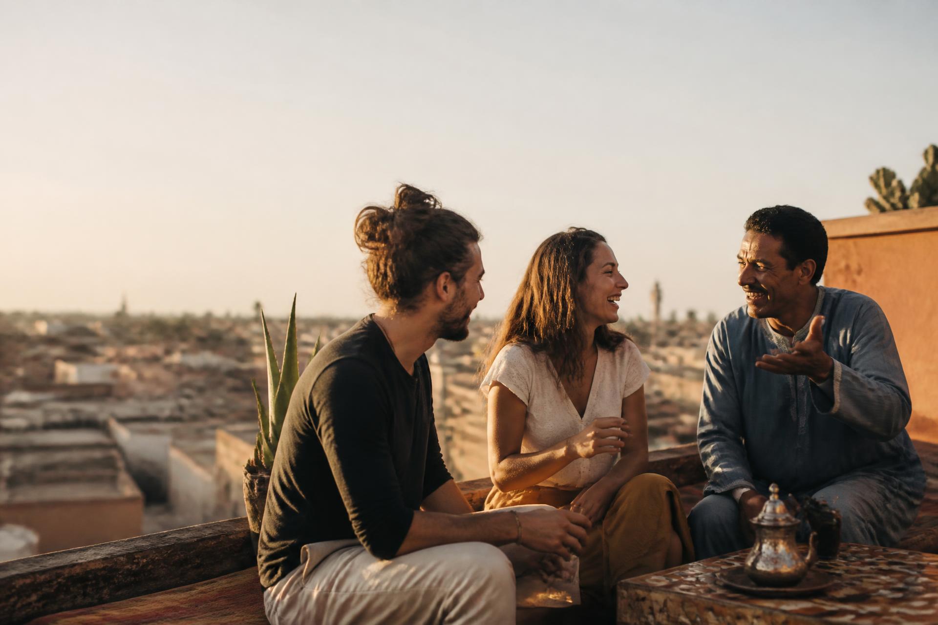 Two travellers in warm conversation with a local host on a sunlit rooftop terrace
